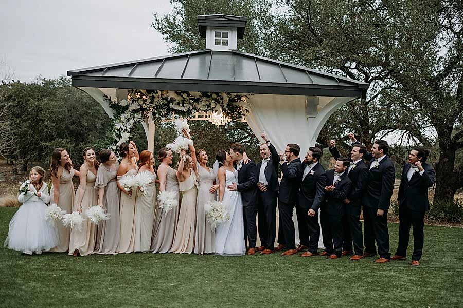 Bride and groom kiss under the gazebo with wedding party looking on.